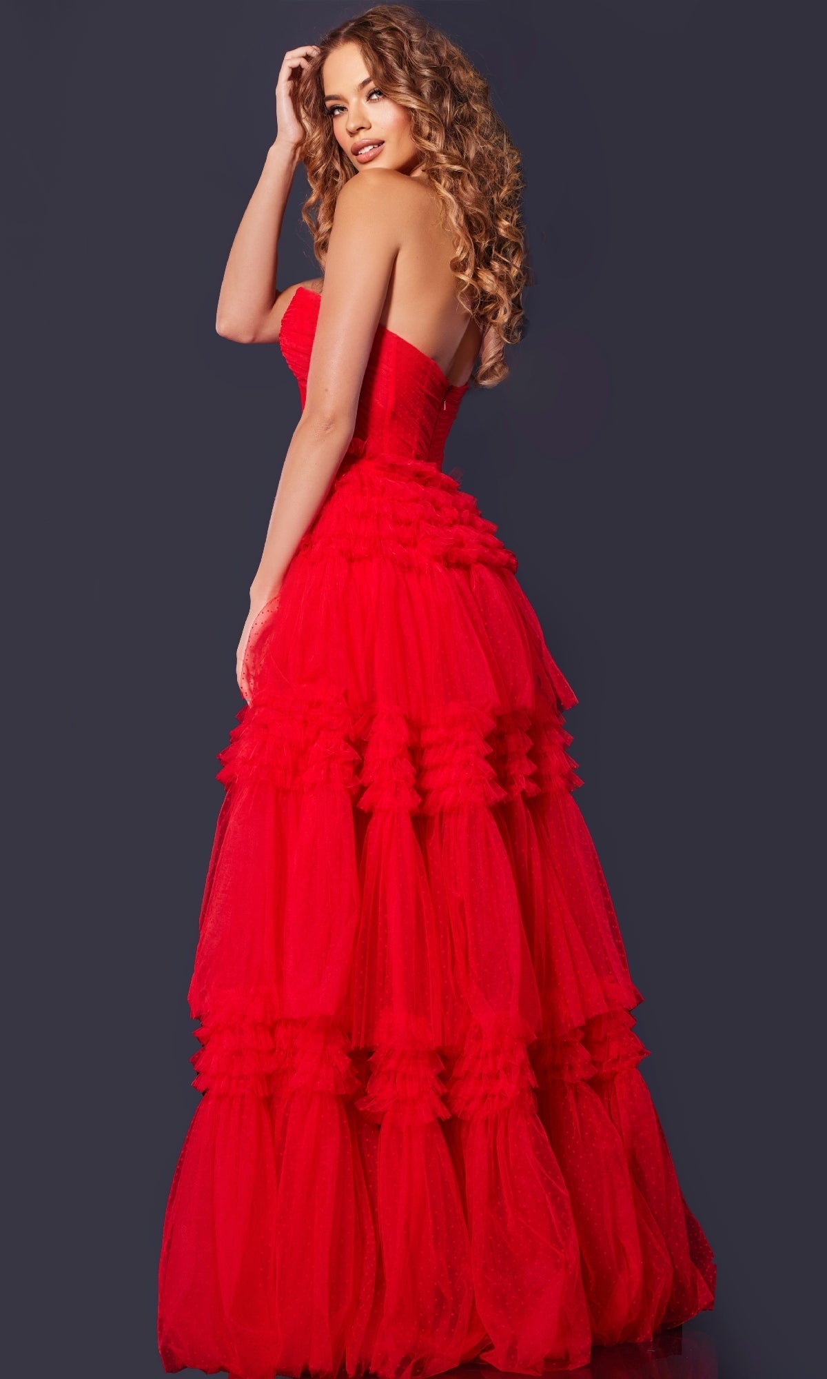 A woman with long curly hair models the Jovani 40466 strapless prom ball gown with a layered tulle skirt, posing against a dark background and looking over her shoulder.
